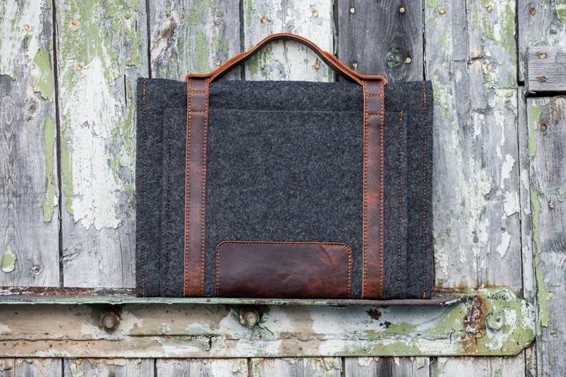 May include: A gray felt laptop sleeve with brown leather accents and a top handle. The sleeve is sitting on a weathered wooden surface.