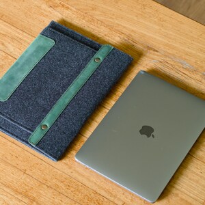Könnte beinhalten: Eine dunkelgraue Laptop-Hülle aus Filz mit grünen Lederakzenten und Riemen, neben einem silbernen Laptop mit dem Apple-Logo. Die Hülle hat einen rechteckigen grünen Aufnäher und einen grünen Riemen.
