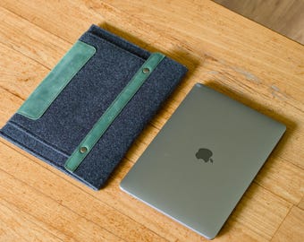 Puede incluir: Una funda para portátil de fieltro gris oscuro con un acento y una correa de cuero verde, junto a un portátil plateado con el logotipo de Apple. La funda tiene un parche rectangular verde y una correa verde.