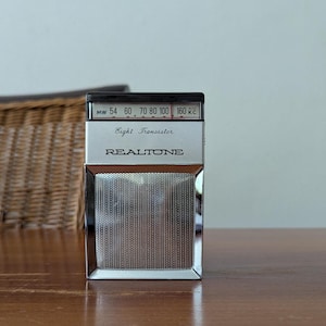 Könnte beinhalten: Ein Vintage RealTone Eight Transistor Radio mit silbernem und schwarzem Finish. Das Radio verfügt über einen Lautsprechergrill, ein Wahlrad und den Text "REALTONE". Das Radio wird auf einer Holzoberfläche angezeigt.