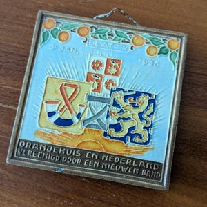 Puede incluir: Un azulejo de cerámica azul y blanco con un escudo de armas holandés, que presenta un león, un cuerno y una cinta. El azulejo está decorado con flores de azahar y el texto "31 JAN. BEATRIX 1938 ORANJEHUIS EN NEDERLAND VERENIGD DOOR EEN NIEUWEN BAND".