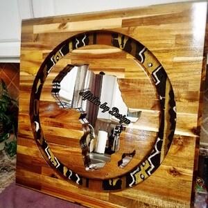 May include: A wooden wall art piece featuring a circular cutout of the continent of Africa. The cutout is filled with a mirror, reflecting the surrounding room. The wood is a light brown color and the mirror is surrounded by a patterned border of black and gold fabric.