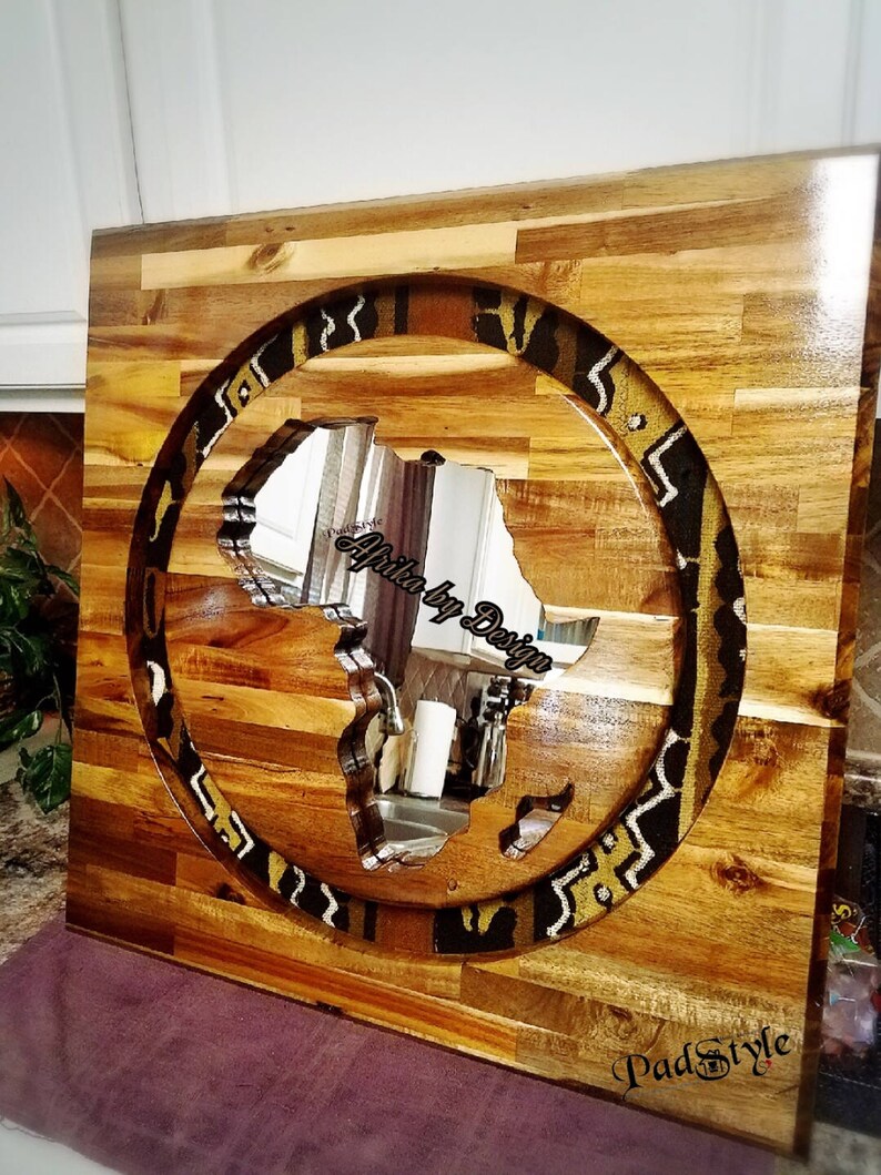 May include: A wooden wall art piece featuring a circular cutout of the continent of Africa. The cutout is filled with a mirror, reflecting the surrounding room. The wood is a light brown color and the mirror is surrounded by a patterned border of black and gold fabric.
