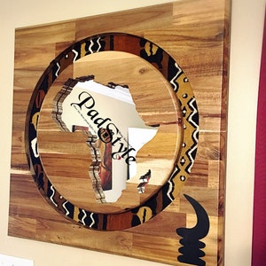 May include: A wooden wall art piece featuring a cutout of the continent of Africa surrounded by a circular patterned border. The border is made of brown, black, and white fabric with geometric designs. The background is a light brown wood.