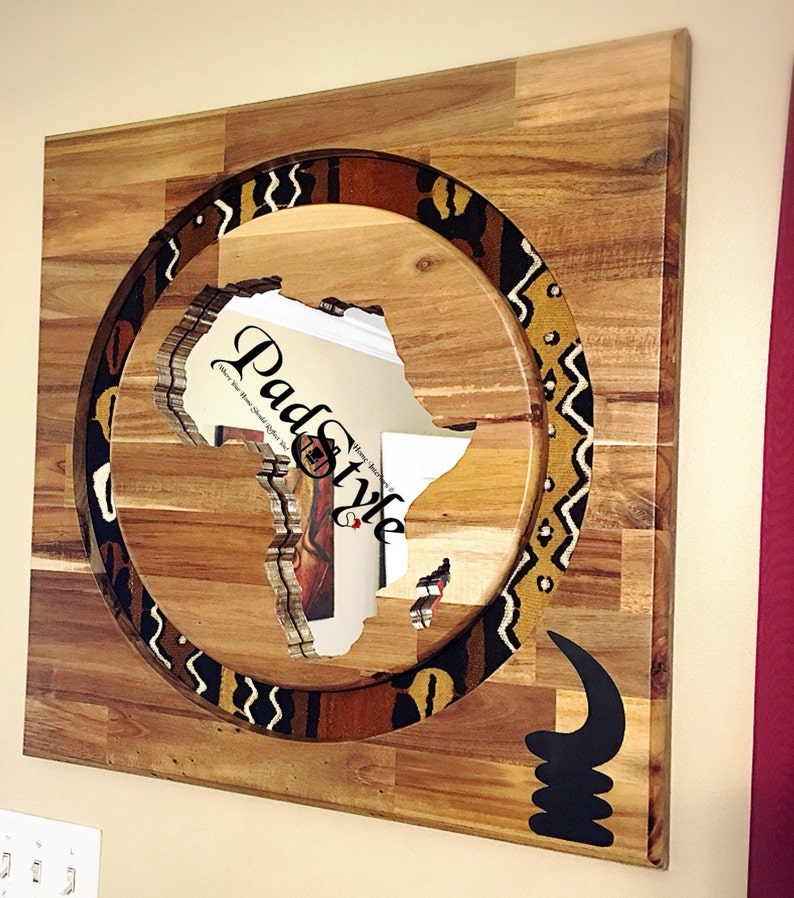 May include: A wooden wall art piece featuring a cutout of the continent of Africa surrounded by a circular patterned border. The border is made of brown, black, and white fabric with geometric designs. The background is a light brown wood.