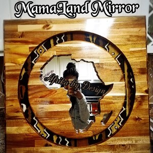 May include: A wooden framed mirror with a circular cutout in the shape of the continent of Africa. The cutout is surrounded by a patterned fabric border. The mirror is labeled "Mama Land Mirror" and "Africa by Design".