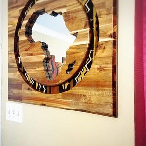 May include: A wooden wall art piece featuring a cutout of the continent of Africa surrounded by a patterned circle. The circle is made of a fabric with black, white, and yellow geometric patterns. The wood is a light brown color.
