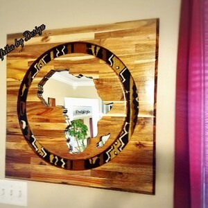 May include: A wooden wall art piece featuring a circular cutout of the continent of Africa with a patterned border. The wood is a light brown color and the pattern is a mix of black, white, and brown.