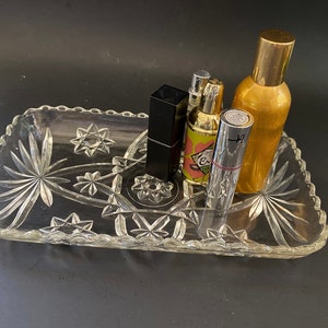 May include: A clear glass tray with a starburst pattern design, holding four bottles of perfume and a lipstick. The bottles are various colors and sizes, including gold, black, and clear.