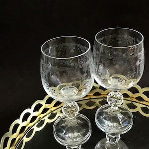 May include: Two clear wine glasses with etched floral designs. The glasses have a round bowl shape and sit on a decorative stem with a faceted base. They are displayed on a gold-colored, scalloped-edge tray against a dark background.