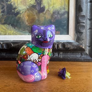 May include: A hand-painted ceramic cat figurine with a purple head and body, featuring green eyes and a red mouth. The cat is decorated with colorful floral and geometric patterns. A small purple flower is next to the cat on a wooden surface.