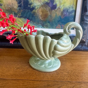 May include: A sage green ceramic vase with a decorative handle and base, holding bright red flowers. The vase has a scalloped rim and sits on a wooden surface, with a framed painting in the background.