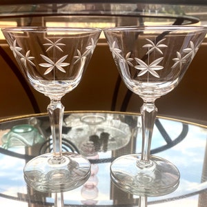 May include: Two clear crystal wine glasses with a starburst design etched into the bowl. The glasses have a long, faceted stem and a round base. The glasses are displayed on a glass-topped table.
