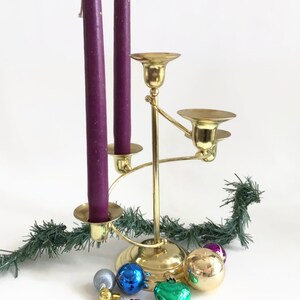 May include: A gold-toned metal candelabra with a central stem and three tiered arms, holding two tall purple taper candles. The base is surrounded by several small, round, multi-colored decorative ornaments and a strand of green artificial pine.