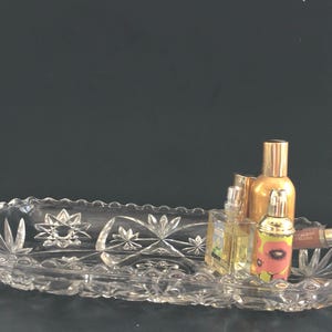 May include: A clear, oval-shaped crystal tray with a scalloped edge, holding several perfume bottles and a tube of lip gloss. The tray features starburst and floral designs. The bottles are gold, clear, and decorated with floral patterns.
