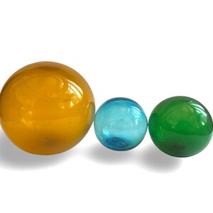 May include: Three glass spheres in a row. The largest is amber, the middle one is light blue, and the smallest is green. The spheres are smooth and reflective, with a slight shadow beneath them.