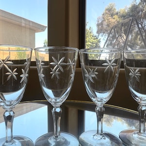 May include: Four clear wine glasses with etched starburst designs. The glasses have a classic shape with a stem and a round base. The background shows a window with a view of trees and a house.
