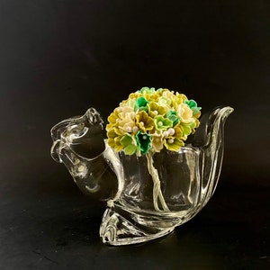 May include: A clear glass vase in a unique shape, holding a bouquet of artificial flowers. The flowers are a mix of green, yellow, and white, with small white bead accents. The vase has a curved design and sits on a dark surface.