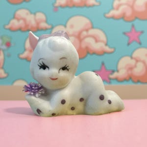 May include: A vintage-style figurine of a reclining cherub with a light green and white finish. The cherub has black eyelashes, a purple flower, and dark purple polka dots. The figurine is on a pink surface.