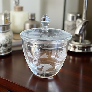 May include: A clear glass jar with a lid, featuring etched leaf and floral designs. The jar has a rounded shape and a small, decorative handle on the lid. The jar is sitting on a dark wooden surface.