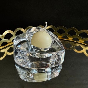 May include: A clear, heart-shaped glass candle holder with a small, round, off-white candle inside. The candle holder sits on a reflective surface, with a gold-colored, scalloped-edge tray in the background. The candle holder is approximately 3 inches wide.
