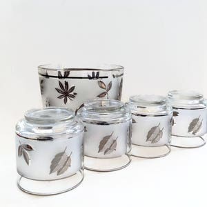 Puede incluir: Un juego de cuatro vasos de vidrio con bordes plateados y una cubitera a juego, todos con un acabado blanco esmerilado y un dise&ntilde;o de hojas plateadas.