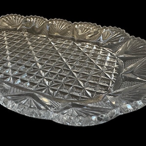 May include: Clear glass serving tray with a diamond pattern in the center and a scalloped edge. The tray has a decorative design around the rim, adding a touch of elegance. This vintage-style tray is perfect for serving appetizers or desserts.