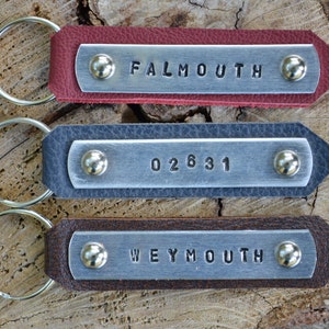 May include: Three leather keychains with silver-toned metal plates attached by two rivets. The top keychain is red with the text FALMOUTH, the middle is dark gray with 02631, and the bottom is brown with WEYMOUTH. Each has a circular metal ring.