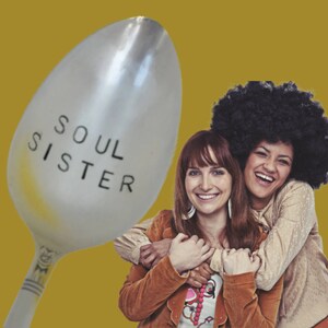 May include: A silver spoon with the words "SOUL SISTER" etched on the bowl. The spoon is set against a mustard-yellow background, with two smiling women in the background. One woman has a large afro.