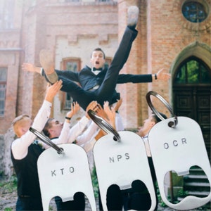 May include: Three white metal bottle openers with rounded rectangular shapes and silver key rings. Each opener features stamped black letters: one reads KTO, the second reads NPS, and the third reads OCR. The openers are displayed in the foreground.