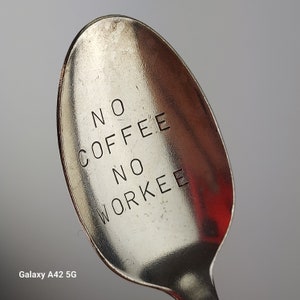 May include: A silver-toned metal spoon featuring the stamped text NO COFFEE NO WORKEE in black capital letters on the bowl. The surface of the spoon shows a metallic sheen with minor surface scratches and a smooth, rounded edge design.