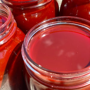 May include: Four glass jars filled with a deep red liquid, likely a homemade fruit preserve or sauce. The jars are arranged in a row, with the top of the jars visible. The liquid is translucent and has a slight sheen.