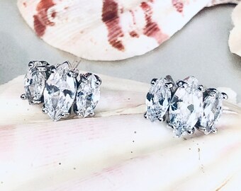 May include: A pair of silver earrings with three marquise-shaped clear stones on each earring. The earrings are displayed on a white seashell.