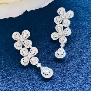 May include: Silver-toned drop earrings in a floral design. Each earring has three flower clusters and a teardrop pendant. The earrings are embellished with clear, sparkling stones. The background is a dark blue, textured surface.