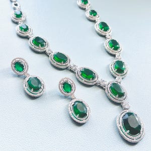 May include: A jewellery set comprising a necklace and earrings. The necklace and earrings feature oval, emerald-green gemstones set in silver-coloured metal. The gemstones are surrounded by a halo of smaller, clear stones.