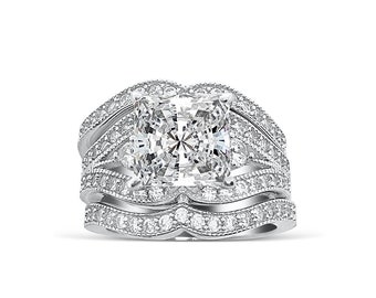 Puede incluir: Un anillo de plata con una gran piedra preciosa cuadrada transparente rodeada de piedras preciosas transparentes más pequeñas. El anillo tiene un diseño delicado e intrincado con múltiples bandas.