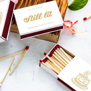 Könnte beinhalten: Weiße Streichholzschachteln mit goldenem Text und weinroten Akzenten sind mit Streichhölzern und floralen Akzenten angeordnet. Auf einer Schachtel steht "Still lit" und eine andere zeigt eine Kuchenillustration. Die Streichhölzer haben rote Spitzen.