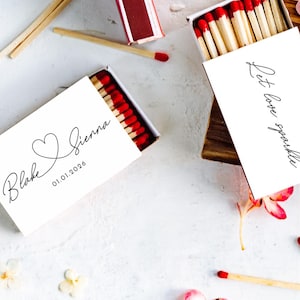 May include: Matchboxes with red-tipped matches, scattered on a white surface with pink and white flowers. One box is open, displaying matches and the text "Blake & Sienna 01.01.2024". Another box has the text "Let love sparkle".