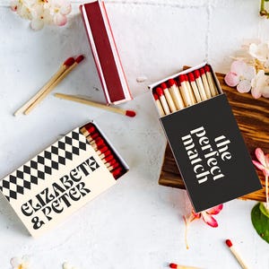 Può includere: Scatole di fiammiferi con fiammiferi dalla punta rossa, sparsi su una superficie bianca con accenti floreali. Una scatola ha un motivo a scacchi in bianco e nero e il testo "ELIZABETH & PETER". Un'altra scatola è nera con "the perfect match".