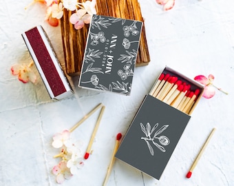 Puede incluir: Dos cajas de cerillas con diseños florales. Una caja tiene el texto "Jill + John 01.01.2024" y la otra caja tiene un diseño floral simple. Ambas cajas son grises con diseños florales blancos.
