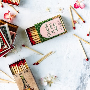 May include: Three matchboxes with different designs. The first matchbox is red and white with the text "THE PAO WOW" and a cartoon image of a woman in a bottle. The second matchbox is green and pink with the text "PARFOIS JE SUIS TRISTE." The third matchbox is red and white with the text "CLASSIC" and a red and white design. There are also several loose matches scattered around the matchboxes.