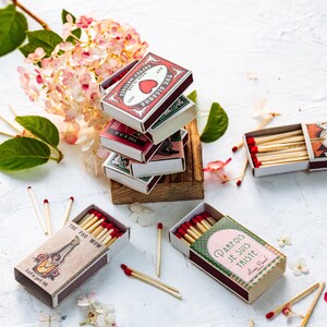 May include: A stack of vintage matchboxes with various designs, including a heart, a dog, and a bottle. The matchboxes are on a wooden surface with pink flowers scattered around. The matchboxes are all closed and have red and white accents.