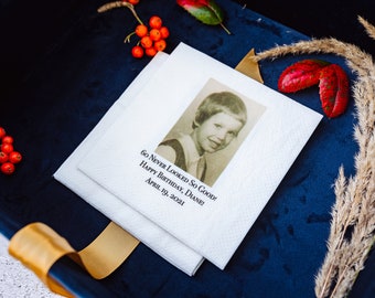 May include: White paper napkins with a vintage photo and text that reads "60 Never Looked So Good! Happy Birthday, Diane! April 19, 2021." The napkins are on a dark blue surface, with decorative elements.