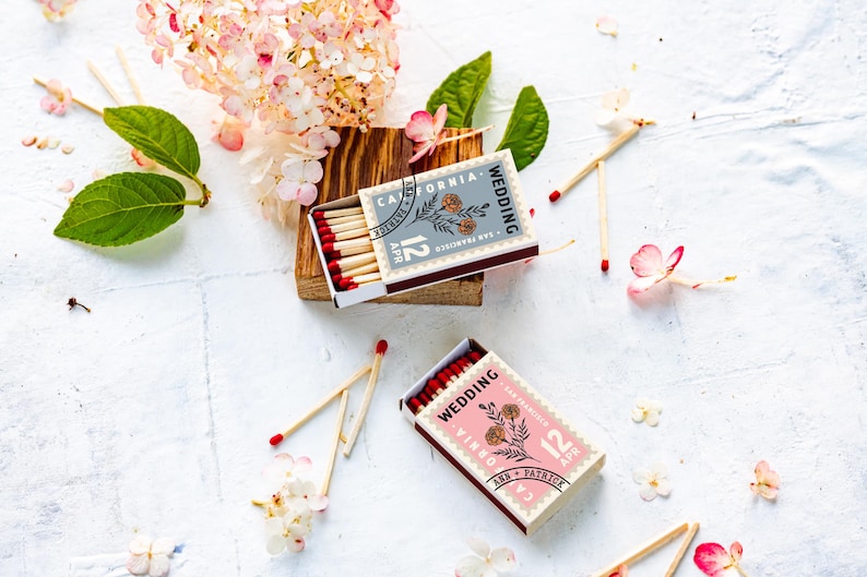 May include: Two rectangular matchboxes featuring a stamp-like design with floral illustrations and text reading Wedding, California, San Francisco, 12 Apr, Ann and Patrick. One box is grey and the other is pink, both containing matches with red tips.