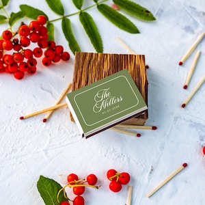 May include: A rectangular matchbox with a sage green cover featuring the white text The Millers and the date 01.07.2020. The matchbox rests on a small wooden block, surrounded by scattered wooden matches and clusters of bright red berries.