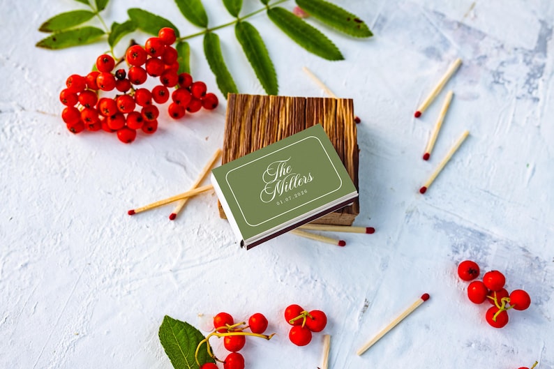 May include: A rectangular matchbox with a sage green cover featuring the white text The Millers and the date 01.07.2020. The matchbox rests on a small wooden block, surrounded by scattered wooden matches and clusters of bright red berries.