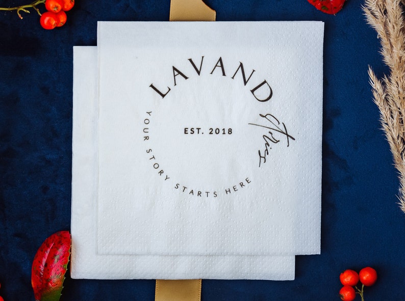 K&ouml;nnte beinhalten: Zwei wei&szlig;e quadratische Servietten mit dem Text "LAVAND" in einem kreisf&ouml;rmigen Design, "EST. 2018" und "YOUR STORY STARTS HERE". Die Servietten liegen auf einer dunkelblauen Oberfl&auml;che mit dekorativen Elementen.