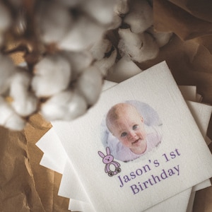 May include: A pile of white square napkins featuring a baby's photo and the text "Jason's 1st Birthday" in purple. A cartoon bunny is next to the text. The napkins are on brown paper, with cotton branches in the background.