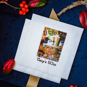 K&ouml;nnte beinhalten: Zwei wei&szlig;e Papierservietten mit einem aufgedruckten Bild einer Frau mit Mickey Mouse-Hut. Der Text "Tracy's 50th" ist darunter gedruckt. Die Servietten liegen auf einer dunkelblauen Oberfl&auml;che, mit roten Beeren und getrockneten Pflanzen.
