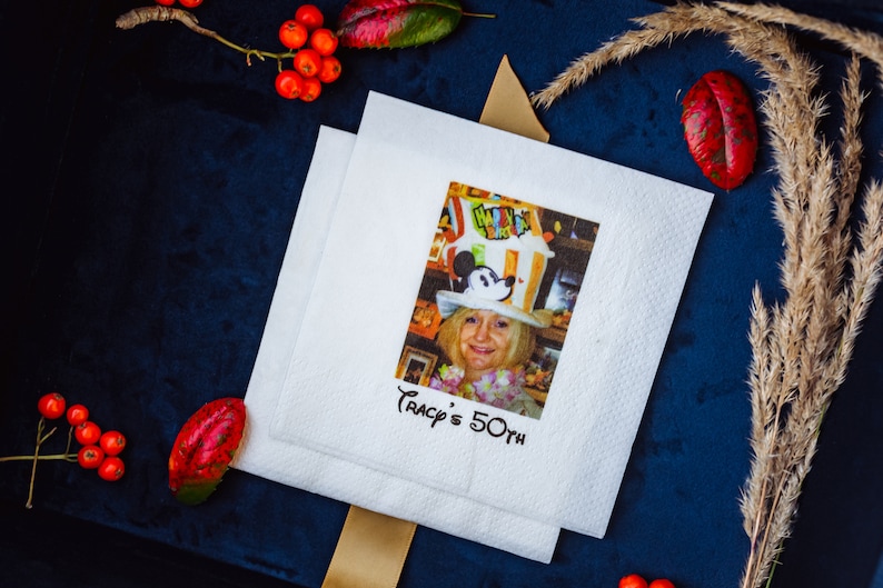 K&ouml;nnte beinhalten: Zwei wei&szlig;e Papierservietten mit einem aufgedruckten Bild einer Frau mit Mickey Mouse-Hut. Der Text "Tracy's 50th" ist darunter gedruckt. Die Servietten liegen auf einer dunkelblauen Oberfl&auml;che, mit roten Beeren und getrockneten Pflanzen.